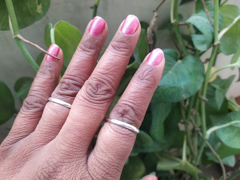 May include: A hand wearing two thin silver rings on the middle and ring fingers. The fingernails are painted a bright pink colour.