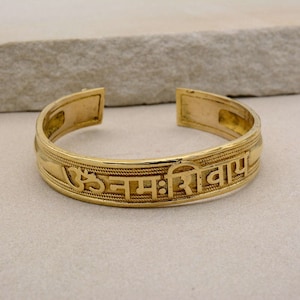 Puede incluir: Un brazalete de color dorado con texto grabado. El brazalete abierto presenta un diseño detallado con una inscripción religiosa. El brazalete se muestra sobre un fondo neutro.