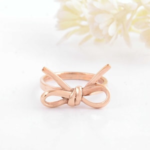 Può includere: Un anello in oro rosa con un design a fiocco. Il fiocco è realizzato in un unico pezzo di metallo ed è annodato.