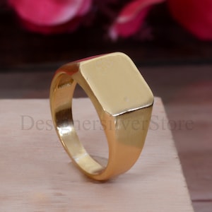 Puede incluir: Un anillo de sello de oro con una parte superior cuadrada. El anillo está hecho de metal sólido y tiene un acabado liso y pulido.