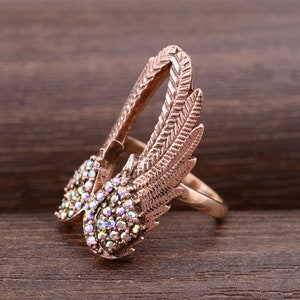 May include: A rose gold ring with a pair of wings adorned with iridescent rhinestones. The wings are detailed with a textured feather design.