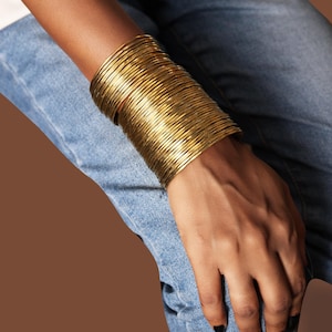 Könnte beinhalten: Ein Stapel goldfarbener Armreifen am Handgelenk. Die Armreifen sind dünn und zylindrisch und bilden einen breiten Armreif. Die Person trägt eine blaue Jeans. Der Hintergrund ist einfarbig braun.