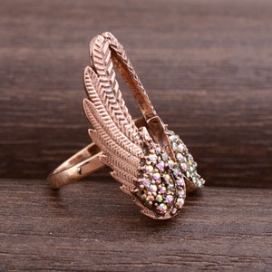 May include: A rose gold ring with a design of two wings with iridescent rhinestones. The wings are detailed with a feather pattern.