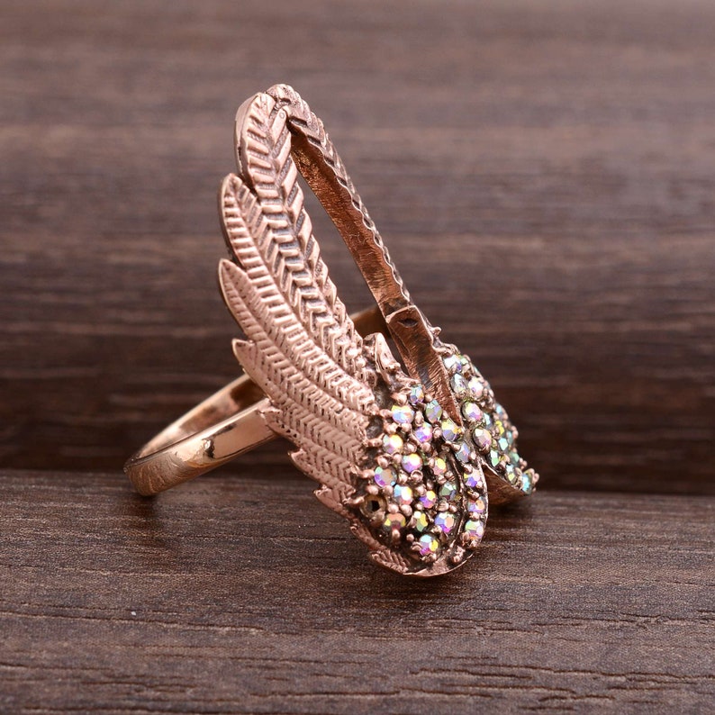 May include: A rose gold ring with a design of two wings with iridescent rhinestones. The wings are detailed with a feather pattern.