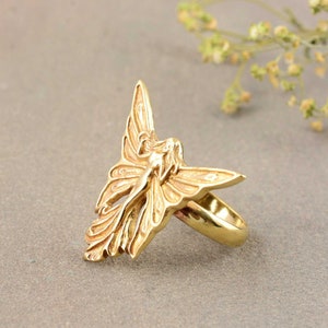 May include: A gold-toned ring featuring a detailed sculpted design of a fairy with wings spread wide.