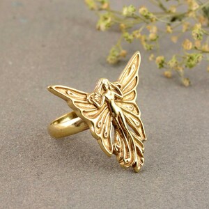 May include: A gold-toned ring featuring a detailed fairy design with wings outstretched. The fairy is depicted with flowing hair and a delicate form.