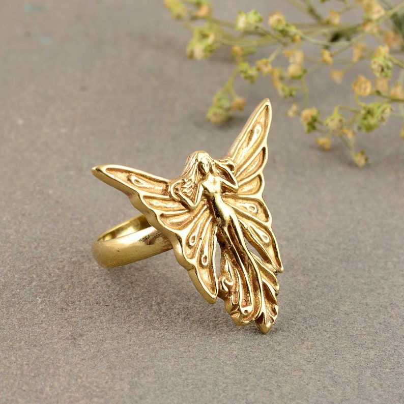 May include: A gold-toned ring featuring a detailed fairy design with wings outstretched. The fairy is depicted with flowing hair and a delicate form.