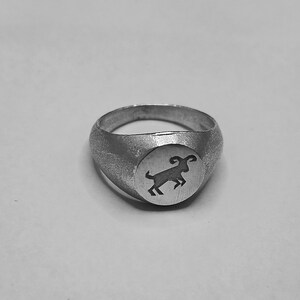 May include: A silver signet ring with a textured band and a circular face. The face displays a black ram silhouette. The ring is a classic design, suitable for everyday wear.