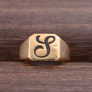 Può includere: Anello con sigillo dorato con la lettera "S" nera in scrittura stilizzata. L'anello ha una faccia rettangolare con bordi smussati. Un gioiello classico, semplice ed elegante, adatto all'uso quotidiano.