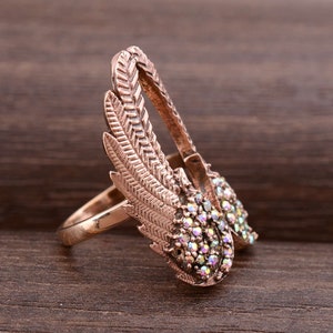 May include: A rose gold ring with a design of two wings with iridescent rhinestones. The wings are detailed with a feather pattern.