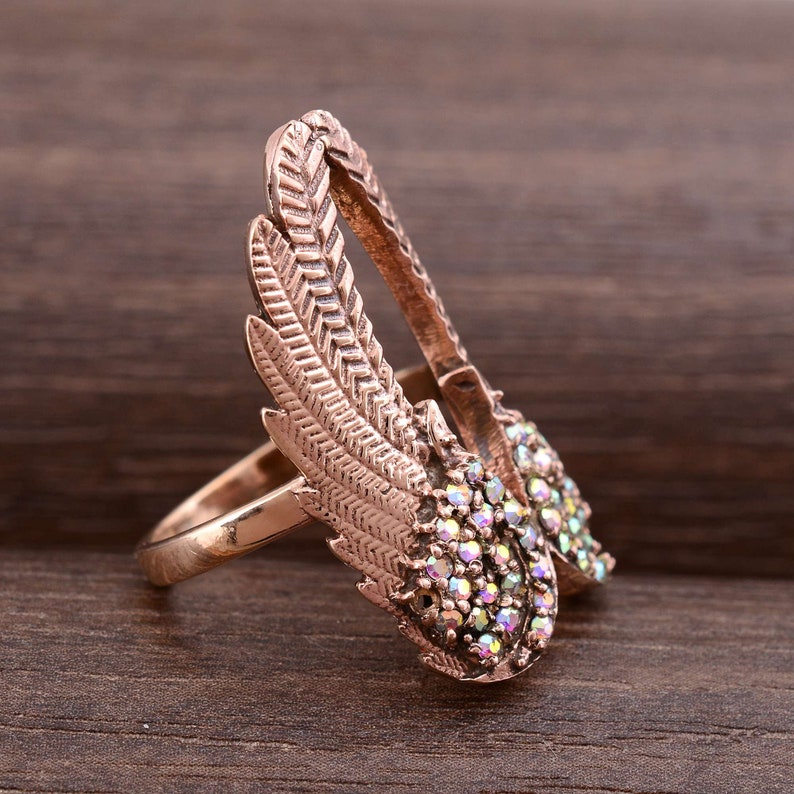 May include: A rose gold ring with a design of two wings with iridescent rhinestones. The wings are detailed with a feather pattern.