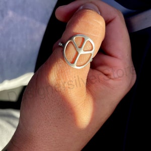 May include: A silver peace sign ring worn on a person's thumb.