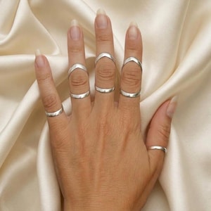 May include: A set of six silver-toned rings worn on the fingers. Three rings feature an open, arched design that sits across the middle knuckle, while three simple, smooth bands are worn at the base of the fingers on the thumb, index, and pinky.