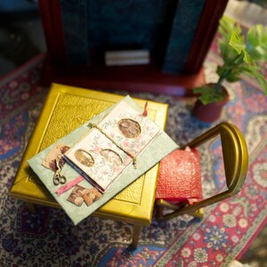 May include: Miniature gold table with a detailed, embossed surface. A small, open photo album with floral accents and framed images rests on top, along with tiny scissors and photos. A red chair and potted plant complete the scene.