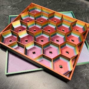 May include: A multi-colored hexagonal tray with 18 compartments. The tray is made of plastic and has a layered, iridescent finish. The tray is open and sitting on top of a smaller, solid purple tray.