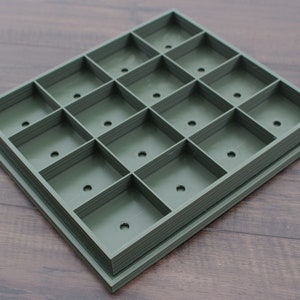 May include: A green plastic tray with 16 square compartments, each with a small hole in the center. The tray is stacked on top of another similar tray.