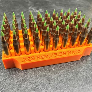 May include: An orange ammunition holder filled with .223 REM./5.56 NATO cartridges. The cartridges have brass casings and green tips. The holder is rectangular and has the caliber information printed on the side.