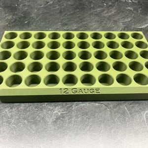 May include: A rectangular olive green storage tray featuring forty-eight circular openings arranged in a grid pattern. The front edge of the tray is embossed with the text 12 GAUGE in capital letters, indicating the intended use for shotgun shells.