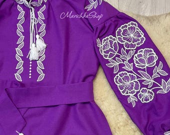 Puede incluir: Un vestido morado vibrante con bordados florales blancos en las mangas y el corpiño. El vestido presenta un diseño de cuello anudado con borlas y un cinturón a juego. El texto "MarichkaShop" es visible.