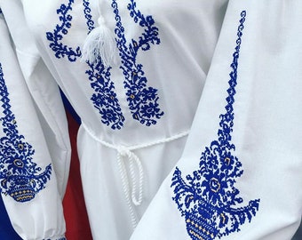 May include: White dress with detailed blue embroidery on the bodice and sleeves. The embroidery showcases floral and geometric designs. A white rope belt defines the waist, and white tassels adorn the neckline. The dress has long sleeves.