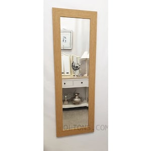 May include: A full-length mirror with a light wood frame. The mirror is reflecting a room with a white dresser and a lamp.