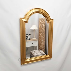 May include: A gold arched mirror with a simple, ornate frame. The mirror is reflecting a white lamp, a small picture frame, and a white dresser with drawers.