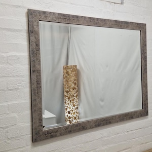Pode incluir: Um espelho retangular com uma moldura texturizada cinza-castanho. O espelho reflete uma sala com uma parede branca e uma lâmpada decorativa. O espelho está montado numa parede de tijolos brancos.