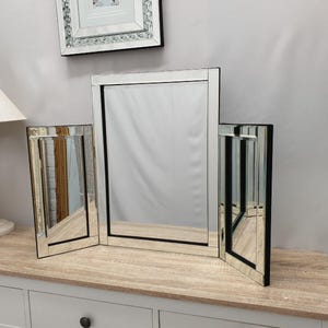 Puede incluir: Un espejo de tocador de tres paneles con un espejo central rectangular y dos paneles laterales. Los espejos tienen un marco plateado con detalles en negro. El espejo está sobre una superficie clara de imitación madera.