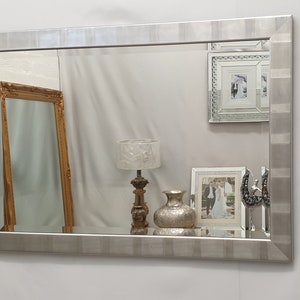 May include: A silver framed mirror with a beveled edge and a textured surface. The mirror is reflecting a room with a gold framed mirror, a lamp with a white shade, a vase, and a framed picture.