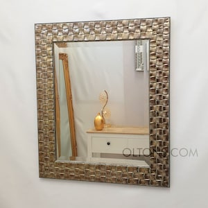 Puede incluir: Un espejo rectangular con un marco texturizado de color dorado. El marco tiene un patrón tejido y un borde ligeramente elevado. El espejo refleja una habitación con una cómoda blanca y un vaso dorado.