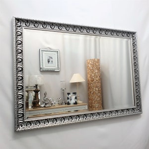 Può includere: Uno specchio grande con cornice argentata decorata con un design ornamentale. Lo specchio riflette una stanza con mobili e decorazioni, tra cui lampade, una cornice per foto e un oggetto decorativo.
