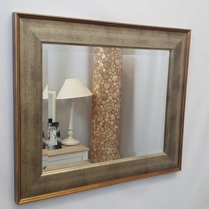 Peut inclure: Un miroir rectangulaire avec un cadre large et orné. Le cadre est de couleur bois vieilli avec une bordure dorée. Le miroir reflète une pièce avec une lampe et un cadre photo.