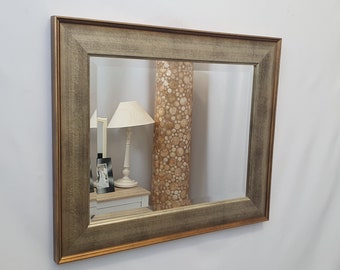 May include: A rectangular mirror with a wide, ornate frame. The frame is a distressed wood color with a gold trim. The mirror is reflecting a room with a lamp and a picture frame.