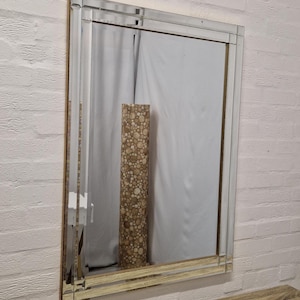 Peut inclure: Miroir rectangulaire avec un bord biseauté, encadré dans un cadre doré. Le miroir reflète une pièce avec un objet cylindrique à motifs et un rideau blanc. Le miroir est monté sur un mur de briques blanches.