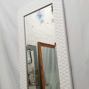 Op de afbeelding: Een witte spiegel in volledige lengte met een getextureerd frame. De spiegel reflecteert een witte bakstenen muur en een andere spiegel met een gouden frame.