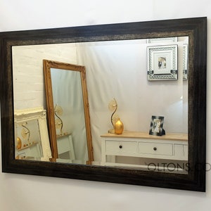 Puede incluir: Un espejo rectangular grande con un marco de madera negro. El marco tiene un acabado desgastado y un borde plateado. El espejo refleja una habitación con muebles y otra decoración.