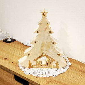 Puede incluir: Adorno navideño de árbol de madera con recortes de estrellas y un pesebre en la base. El árbol está colocado sobre un mantelito de encaje blanco y sobre una superficie de madera. El árbol está iluminado.