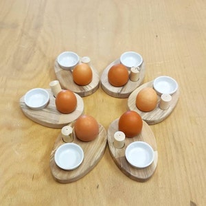 May include: Six wooden egg holders, each with a brown egg, a small white bowl, and a salt shaker. The holders are arranged in a circular pattern on a light brown wooden surface.