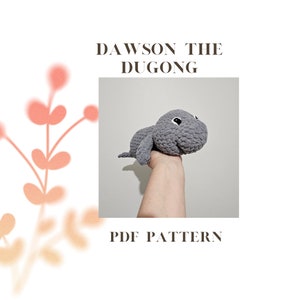 May include: A crocheted gray dugong plush toy with black eyes. The toy is being held by a hand. The text "DAWSON THE DUGONG PDF PATTERN" is below the image.