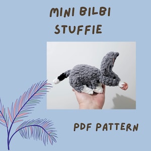 Könnte beinhalten: Ein graues und weißes gehäkeltes Bilby-Plüschtier mit einem schwarzen Schwanz. Das Plüschtier sitzt auf einer Hand. Der Text "MINI BILBI STUFFIE PDF PATTERN" befindet sich unter dem Bild.