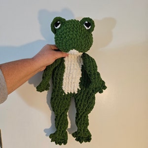 Puede incluir: Un juguete de peluche de rana verde tejido a crochet con detalles de hilo blanco. La rana tiene grandes ojos negros y una amplia sonrisa.