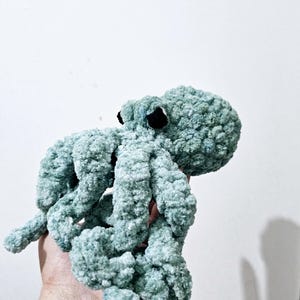 Può includere: Un giocattolo di peluche a forma di polpo all'uncinetto di colore blu-verde chiaro. Il polpo ha otto tentacoli e occhi neri.