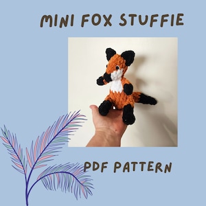 May include: A crocheted fox stuffed animal with orange and brown fur, black ears and nose, and a white chest. The fox is sitting on a hand, and the image includes the text "Mini Fox Stuffie PDF Pattern".