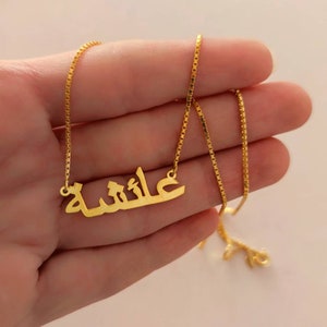 Puede incluir: Un collar de cadena dorada con un colgante que presenta la palabra árabe "عائشة" en una fuente simple y moderna.
