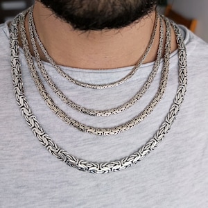 May include: Four silver chain necklaces of varying lengths and widths, layered on a person's neck. The chains are all made of a woven pattern.