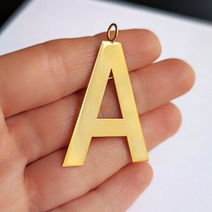 Könnte beinhalten: Ein goldfarbener Anhänger aus Metall in Form des Buchstabens A. Der Buchstabe ist ausgeschnitten und hat eine glatte, glänzende Oberfläche.