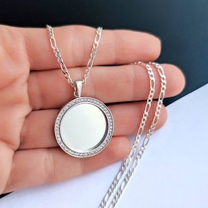 May include: A silver chain necklace with a round pendant featuring a clear, circular, glass-like insert surrounded by small, clear stones.