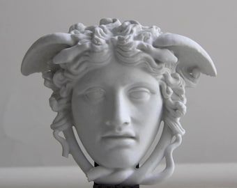 May include: A white sculpture of a woman's face, possibly Medusa, with detailed curly hair and wings. The sculpture is mounted on a dark brown rectangular base. The face has a serene expression.
