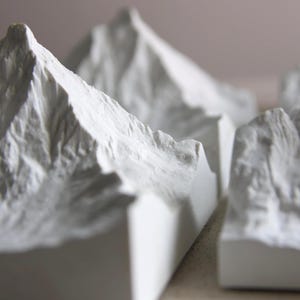 May include: A series of white, miniature mountain sculptures with detailed, textured surfaces. The peaks and ridges are sharply defined, creating a realistic landscape. The sculptures are arranged in a row, showcasing the varying heights and shapes of the mountains.