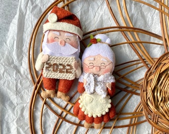 Puede incluir: Adornos navideños de fieltro hechos a mano con Papá Noel y Mamá Noel. Papá Noel sostiene un letrero con "2022". Ambas figuras tienen mejillas rosadas y visten trajes festivos en tonos rojos, blancos y beige. Los adornos están sobre un fondo tejido.